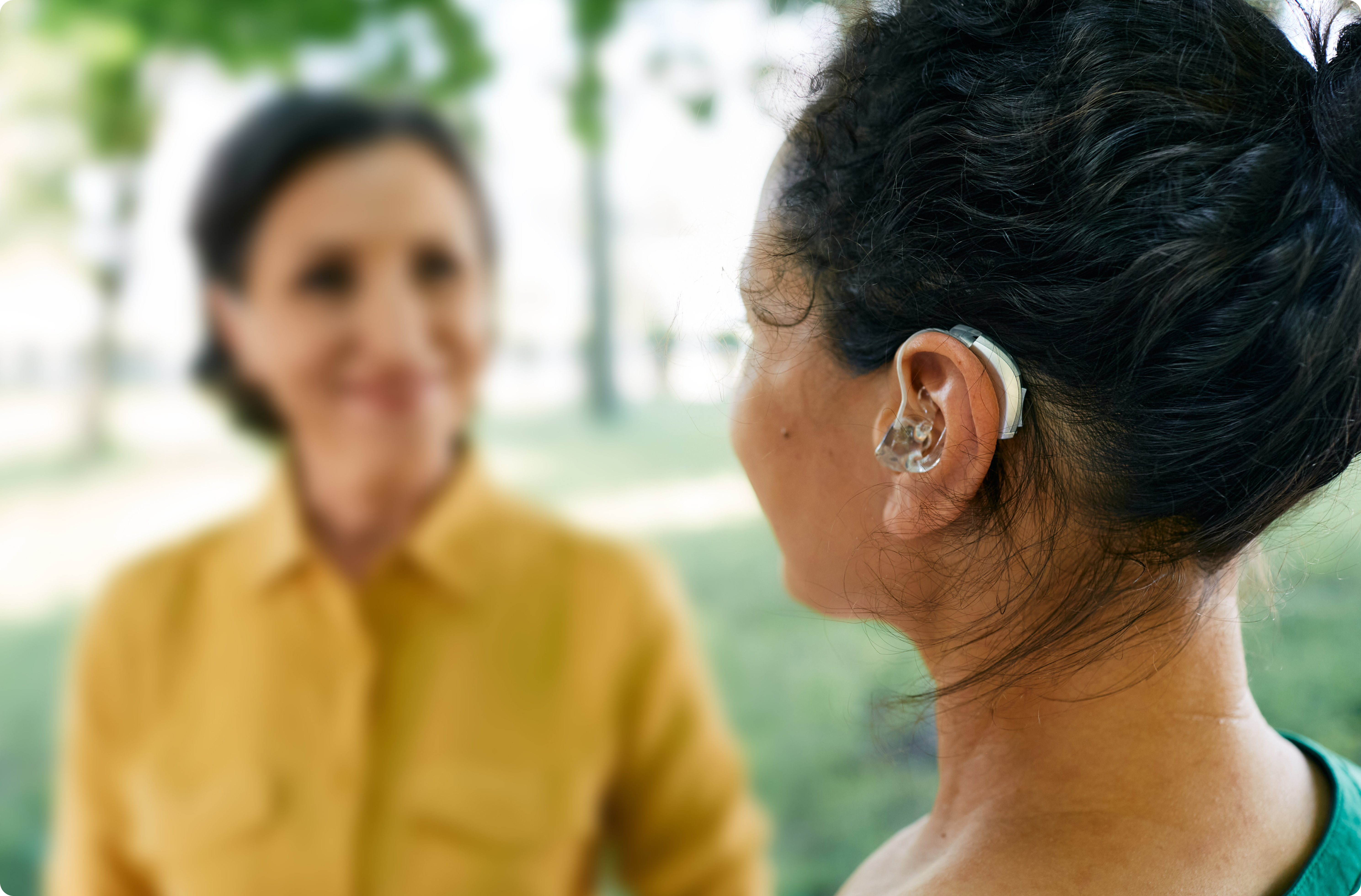 Picture of a young woman wearing a hearing aid, while talking to an older woman in blurred view. 