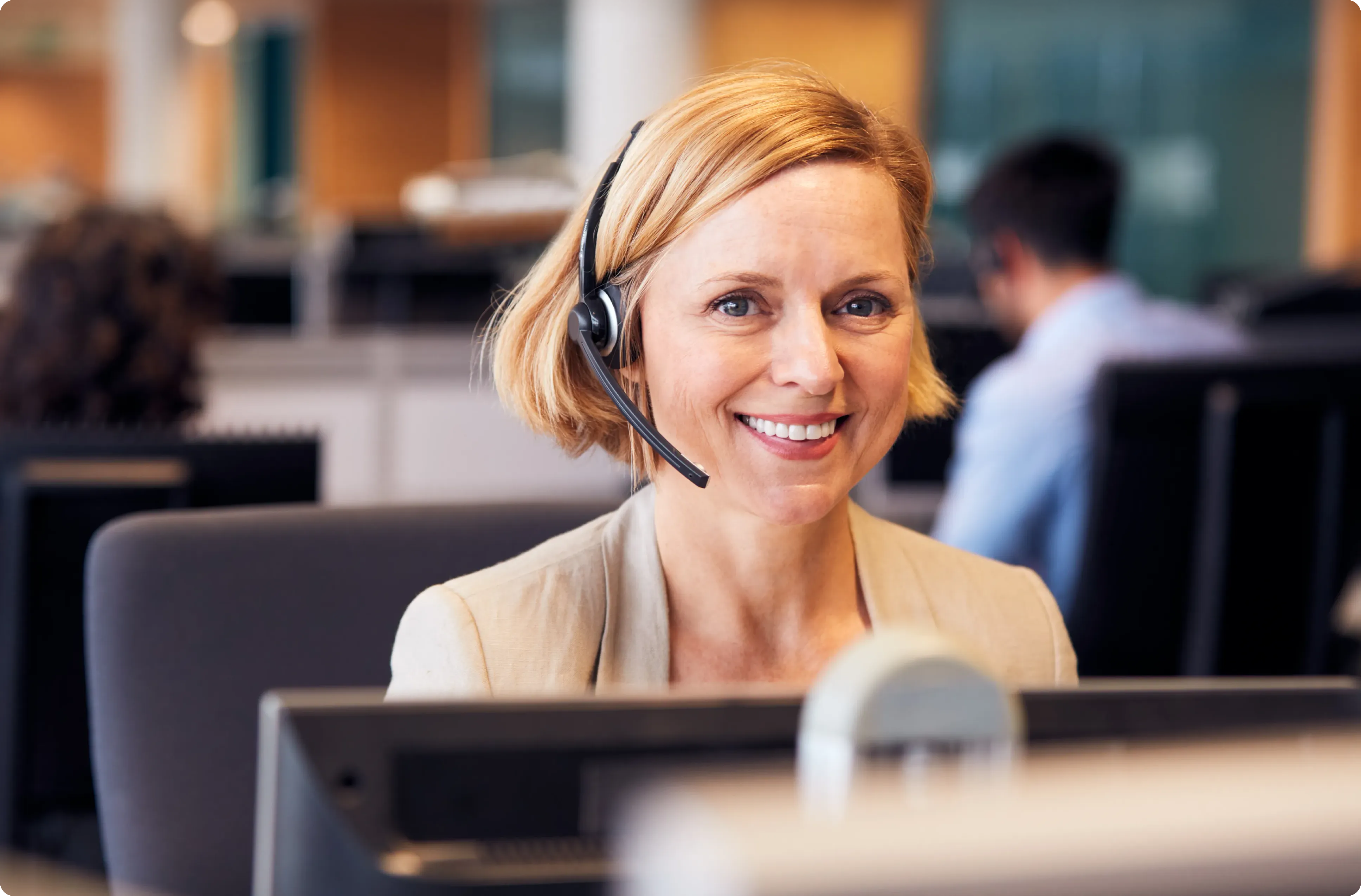 Picture of a customer service operator wearing a headset in an open plan office, smiling at the camera.